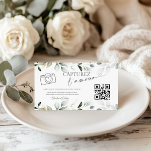 Puede incluir: Una tarjeta blanca con el texto "CAPTUREZ l'amour" y una ilustración de cámara, rodeada de hojas verdes. La tarjeta está sobre un plato blanco, con rosas blancas y vegetación en el fondo. La tarjeta también tiene un código QR.
