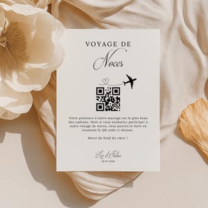 Peut inclure: Une carte blanche avec le texte "Voyage de Noces" en lettres noires. Un code QR se trouve au centre de la carte avec un petit avion qui vole au-dessus. Le texte sous le code QR se lit comme suit : "Votre présence à notre mariage est le plus beau des cadeaux. Mais si vous souhaitez participer à notre voyage de noces, vous pouvez le faire en scannant le QR code ci-dessus. Merci du fond du cœur !" Les noms "Lar & Julion" et la date "28.07.2040" sont en bas de la carte.