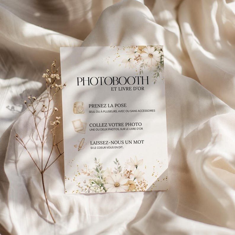 May include: A rectangular sign with the text "PHOTOBOOTH ET LIVRE D'OR" in black. The sign has floral and gold accents. The sign includes instructions in French: "PRENEZ LA POSE", "COLLEZ VOTRE PHOTO", and "LAISSEZ-NOUS UN MOT".