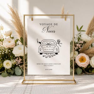 Op de afbeelding: Een goudkleurig metalen frame met een crèmekleurige papieren inleg. De inleg bevat een zwart-wit illustratie van een auto met de woorden "VOYAGE DE Noces" en "LEA & JULIEN". De tekst "MERCI DE NOUS AIDER À BOUCLER NOS VALISES!" is ook aanwezig.