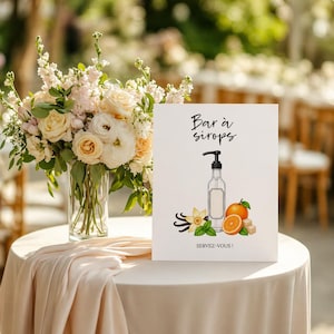 Peut inclure: Une pancarte blanche avec le texte "Bar à sirops" et "SERVEZ-VOUS!" présente une illustration de bouteille de sirop avec des tranches d'orange, des gousses de vanille et des morceaux de sucre. Un bouquet de fleurs est posé dans un vase en verre sur une nappe blanche.