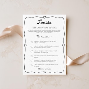 Könnte beinhalten: Eine weiße, rechteckige Hochzeitstischkarte mit einem schwarzen, dekorativen Rand, der mit kleinen Herzen verziert ist. Der Text lautet Louise, Tu es la capitaine de table, und listet verschiedene Hochzeitsaufgaben auf, signiert mit Olivia & Damien.