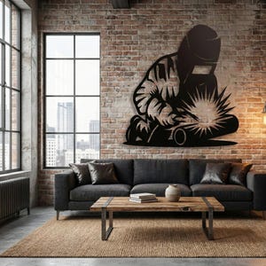 May include: Black metal wall art depicting a welder at work, with a bright burst of light emanating from the welding torch. The artwork is mounted on a brick wall above a dark gray sofa in a modern living room setting.
