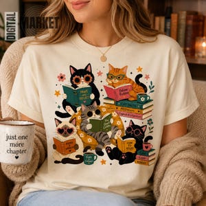 May include: A cream-coloured t-shirt featuring a graphic of five cats reading books. The cats wear glasses and are surrounded by stacks of books with titles like Paws and Reflect, Feline Fiction, and Meowoirs. A white mug with the text just one more chapter is held in the foreground.
