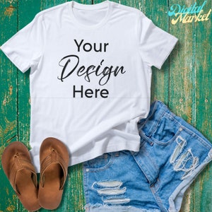 Puede incluir: Camiseta blanca con el texto "Your Design Here" sobre un fondo de madera verde. La camiseta est&aacute; sobre un par de pantalones cortos de mezclilla azul y chanclas marrones.