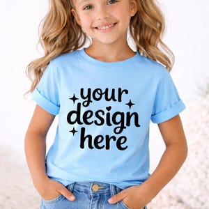 May include: A light blue t-shirt with rolled-up sleeves featuring the text "your design here" in black. The shirt is worn by a young girl with blonde hair and denim shorts. Pink bows adorn her pigtails.