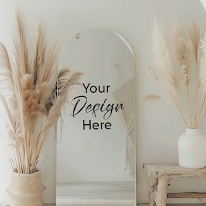以下が含まれることがあります： 白い壁に大きなアーチ型の鏡がかかっています。鏡には、白い部屋に木の踏み台と白い花瓶に生けた乾燥したパンパスグラスが映っています。鏡には「Your Design Here」という文字が書かれています。