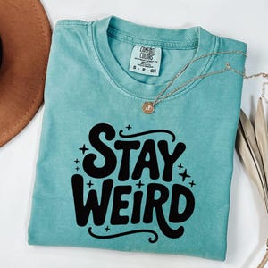 Peut inclure: Un t-shirt turquoise Comfort Colors avec l'inscription "STAY WEIRD" en lettres noires grasses. Le t-shirt a un col rond et des manches courtes. Un collier en or avec un pendentif est drapé sur le t-shirt.