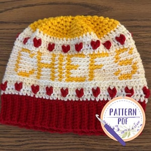 May include: A crocheted hat with a red, white, and yellow color scheme. The hat has a heart pattern and the word "CHEER" is crocheted into the design. The hat is accompanied by a sign that says "PATTERN PDF".