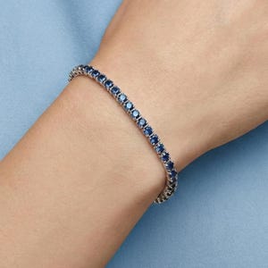 Peut inclure: Un bracelet en argent orné d'une rangée continue de pierres précieuses rondes bleu foncé. Le bracelet est porté sur un poignet sur fond bleu clair. Les pierres sont espacées et serties dans une monture argentée.