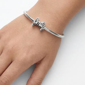 Peut inclure: Un bracelet argenté avec un motif de chaîne serpent. Le bracelet porte le mot "family" en écriture cursive comme breloque. Le bracelet est porté au poignet.