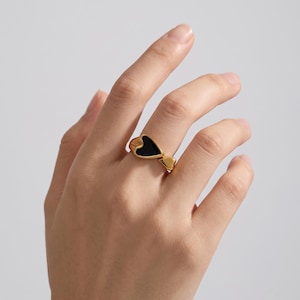 Peut inclure: Une bague dorée présentant un motif en forme de cœur avec une incrustation noire lisse. L'anneau est poli et métallique, mettant en valeur un élément secondaire en forme de petit cœur adjacent au cœur noir principal.