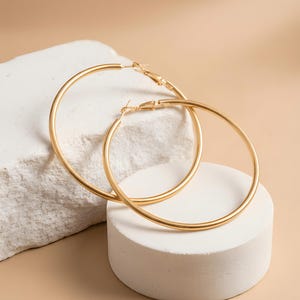 Peut inclure: Une paire de grandes créoles dorées, fines et circulaires, dotées d'une finition lisse et polie. Les boucles d'oreilles présentent un mécanisme de fermeture simple par tige sur le dessus, conçu pour être porté au lobe de l'oreille.