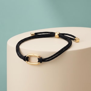 Op de afbeelding: Een zwart koordarmband met een centrale gepolijste goudkleurige ovale schakel. De armband is voorzien van een verstelbaar schuifmechanisme met een goudkleurige kraal en twee kleine goudkleurige eindkapjes aan de uiteinden van het koord.