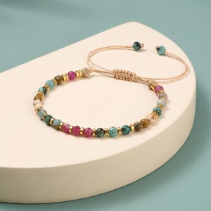 Peut inclure: Un bracelet de perles composé de perles rondes à facettes dans des tons turquoise, magenta et beige, alternées avec de petites entretoises cylindriques dorées. Le bracelet possède un cordon beige avec un fermoir coulissant réglable.