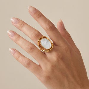 Puede incluir: Un anillo de tono dorado con una incrustación central ovalada de nácar blanco. El anillo presenta un diseño de marco abierto con un mecanismo giratorio que permite que la pieza central ovalada gire dentro de la banda metálica dorada.