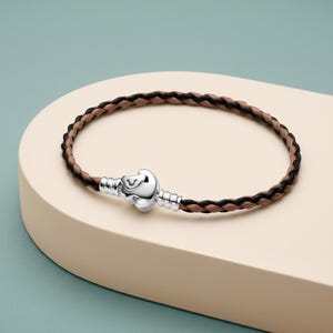 Puede incluir: Una pulsera trenzada con hilos alternos de cuero negro y marrón claro. La pulsera se asegura con un cierre de metal plateado pulido que presenta una forma redondeada y lisa, con un diseño estriado en los extremos de conexión.
