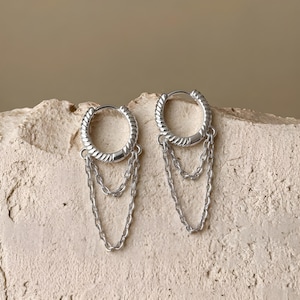 Peut inclure: Une paire de boucles d'oreilles créoles argentées présentant une texture métallique torsadée sur les anneaux. Chaque créole est ornée de deux fines chaînes argentées pendantes qui forment une courbe superposée sous l'anneau circulaire.