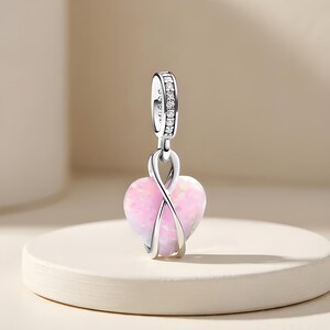 Pode incluir: Um berloque de prata com uma pedra de opala rosa em forma de coração, envolta numa armação de prata polida com design de infinito. O passador superior é decorado com pequenas pedras transparentes, servindo de fixação para pulseiras ou correntes.