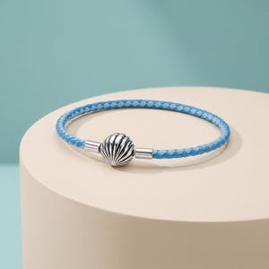 Peut inclure: Un bracelet tressé composé d'un cordon tissé bleu clair et blanc. Le bracelet est maintenu par un fermoir en métal argenté poli en forme de coquillage arrondi avec des stries verticales, assurant une fermeture sécurisée pour cet accessoire.