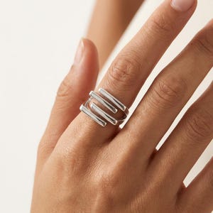 May include: A polished silver ring featuring a multi-layered design with three distinct, parallel horizontal bars that wrap around the finger. The metal has a smooth, reflective finish and a structured, open-ended band construction.