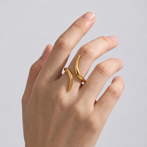 May include: A gold-toned open ring featuring two curved, tapered ends that extend across the finger. The polished metal surface has a smooth, reflective finish, creating a continuous, fluid shape that wraps partially around the finger.