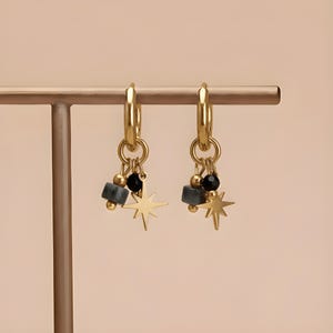 May include: A pair of gold hoop earrings featuring dangling charms. Each earring includes a small gold star, a smooth black bead, a small gold sphere, and a grey cylindrical bead suspended from a circular gold ring attached to the main hoop.