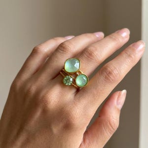 Peut inclure: Une bague dorée ornée de trois pierres vertes distinctes. Le design comprend une grande pierre vert clair de taille carrée, une petite pierre vert clair de taille ronde et une pierre vert clair moyenne de taille ronde, sur un anneau doré texturé.