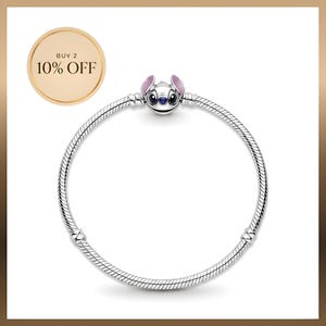 Peut inclure: Un bracelet en chaîne serpent argentée avec un charm représentant le personnage Stitch, doté de grands yeux noirs, d'un nez bleu et d'oreilles roses. Un badge doré circulaire en haut à gauche affiche le texte BUY 2 10% OFF en noir.