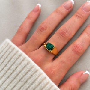May include: A gold-toned ring featuring a smooth, polished band and a central, bezel-set malachite gemstone. The stone is oval-shaped with distinct dark green banding, set within a sleek, minimalist metal frame that sits flush against the finger.