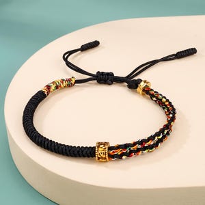 Peut inclure: Un bracelet en cordon noir doté d'une perle centrale en métal doré avec des symboles gravés. Un côté du bracelet est un cordon tressé noir uni, tandis que l'autre côté se compose de fils multicolores en rouge, jaune et vert.