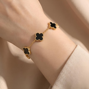 Può includere: Un bracciale a catena dorata con quattro ciondoli neri a forma di quadrifoglio. Ogni ciondolo presenta un bordo dorato testurizzato ed è collegato da una sottile catena a maglie dorate. Il bracciale è indossato al polso, mostrando il motivo.