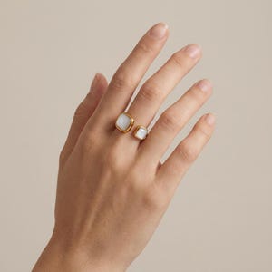 Peut inclure: Une bague ouverte dorée ornée de deux pierres blanches carrées de tailles différentes. La plus grande pierre est placée à une extrémité de l'anneau, tandis que la plus petite se trouve à l'autre extrémité, créant un espace entre les deux.