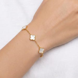Peut inclure: Un bracelet doré orné de quatre motifs en forme de trèfle blanc. Le bracelet est porté au poignet, mettant en valeur le contraste entre la chaîne dorée et les motifs blancs. Un bijou élégant, idéal pour toutes les occasions.