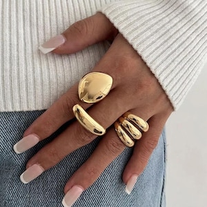 Peut inclure: Un ensemble de trois bagues dorées portées aux doigts. Une bague présente un grand design lisse et irrégulier en forme de galet. Une autre est un anneau épais et incurvé. La troisième bague se compose de trois anneaux polis, arrondis et empilés.