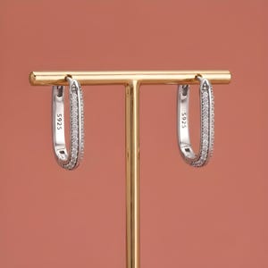 May include: A pair of silver-toned oval hoop earrings featuring a row of small clear stones along the front edge. Each earring is stamped with the text S925 on the inner side. The earrings are displayed hanging from a gold-toned metal T-bar stand.