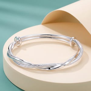 Peut inclure: Un bracelet jonc argenté au design multi-brins composé de deux bandes entrelacées. Une bande présente une finition lisse et polie, tandis que l'autre bande affiche une surface texturée et scintillante. Le bracelet comprend deux petits arrêts.