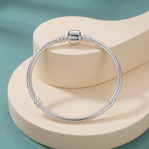 Puede incluir: Una pulsera de cadena de serpiente plateada con un cierre cilíndrico grabado con el texto S925. La pulsera tiene un diseño liso y flexible con dos estaciones fijas a lo largo de la cadena, proporcionando un cierre seguro para esta pieza de joyería.