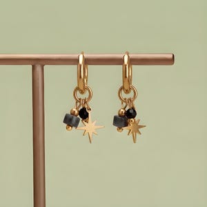 May include: A pair of gold hoop earrings featuring dangling charms. Each earring includes a small gold star, a smooth black bead, and a grey cylindrical bead suspended from a gold ring attached to the main hoop, creating a layered design.