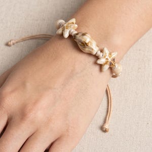 Peut inclure: Un bracelet composé de deux perles en forme d'étoile de mer couleur crème et d'une perle centrale en forme de coquillage spiralé, montées sur un cordon beige clair. Le design inclut de petites perles rondes entre les éléments en coquillage.
