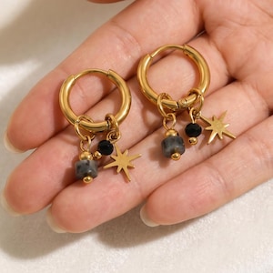 May include: A pair of gold-toned hoop earrings featuring dangling charms. Each hoop suspends a small eight-pointed star, a round black bead, and a cylindrical grey stone bead, all attached with small gold-toned jump rings and metal accents.