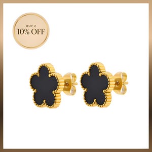 Op de afbeelding: Een paar goudkleurige oorknopjes met een zwart, vierbladig bloemmotief en een kralenrand. Een cirkelvormige gouden badge in de linkerbovenhoek toont de tekst BUY 2 10% OFF in zwarte letters tegen een lichtgouden achtergrond.