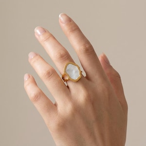 Peut inclure: Une bague dorée dotée d'un sertissage de forme organique irrégulière avec une incrustation nacrée blanche et lisse. L'anneau en métal présente une texture perlée délicate sur le bord extérieur du sertissage, créant un contraste marqué.