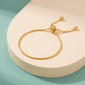 May include: A gold-toned snake chain bracelet featuring an adjustable sliding mechanism. The bracelet consists of a flexible, smooth metallic cord that forms a circular loop, finished with two small spherical beads at the ends of the pull strings.