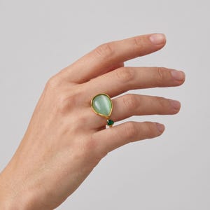 Peut inclure: Une bague en or ornée d'une grande pierre lisse en forme de goutte d'un vert pâle et d'une plus petite pierre triangulaire d'un vert émeraude profond. L'anneau est fin et métallique, avec une finition dorée polie qui met en valeur les deux gemmes.