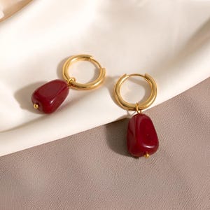 Peut inclure: Une paire de boucles d'oreilles créoles dorées, chacune dotée d'une breloque en forme de galet, lisse et rouge foncé, suspendue à la base. Les breloques présentent une finition polie et brillante, fixées aux anneaux par de petits embouts dorés.