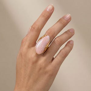 May include: A gold ring featuring a large, smooth, oval-shaped light pink stone set in a polished gold band. The setting curves around the sides of the gemstone, which has a soft, opaque appearance and a subtle, marbled texture across its surface.