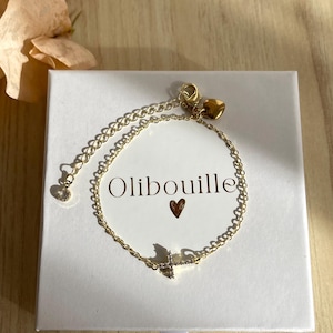 Peut inclure: Un bracelet doré avec une chaîne délicate et un petit pendentif en forme de croix étincelante. Le bracelet est présenté sur une boîte blanche avec le mot "Olibouille" et un symbole de cœur. Le fond est une surface en bois clair.