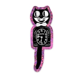 May include: A black cat-shaped wall clock with a white bow tie and a pink glitter border. The clock face is black with white numbers and hands. The text "Lil Cat" is visible on the clock face.