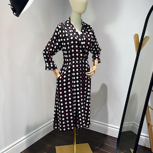 May include: A black and white polka dot dress with red accents. The dress has a collared neckline and three-quarter sleeves. The dress is on a gold stand.